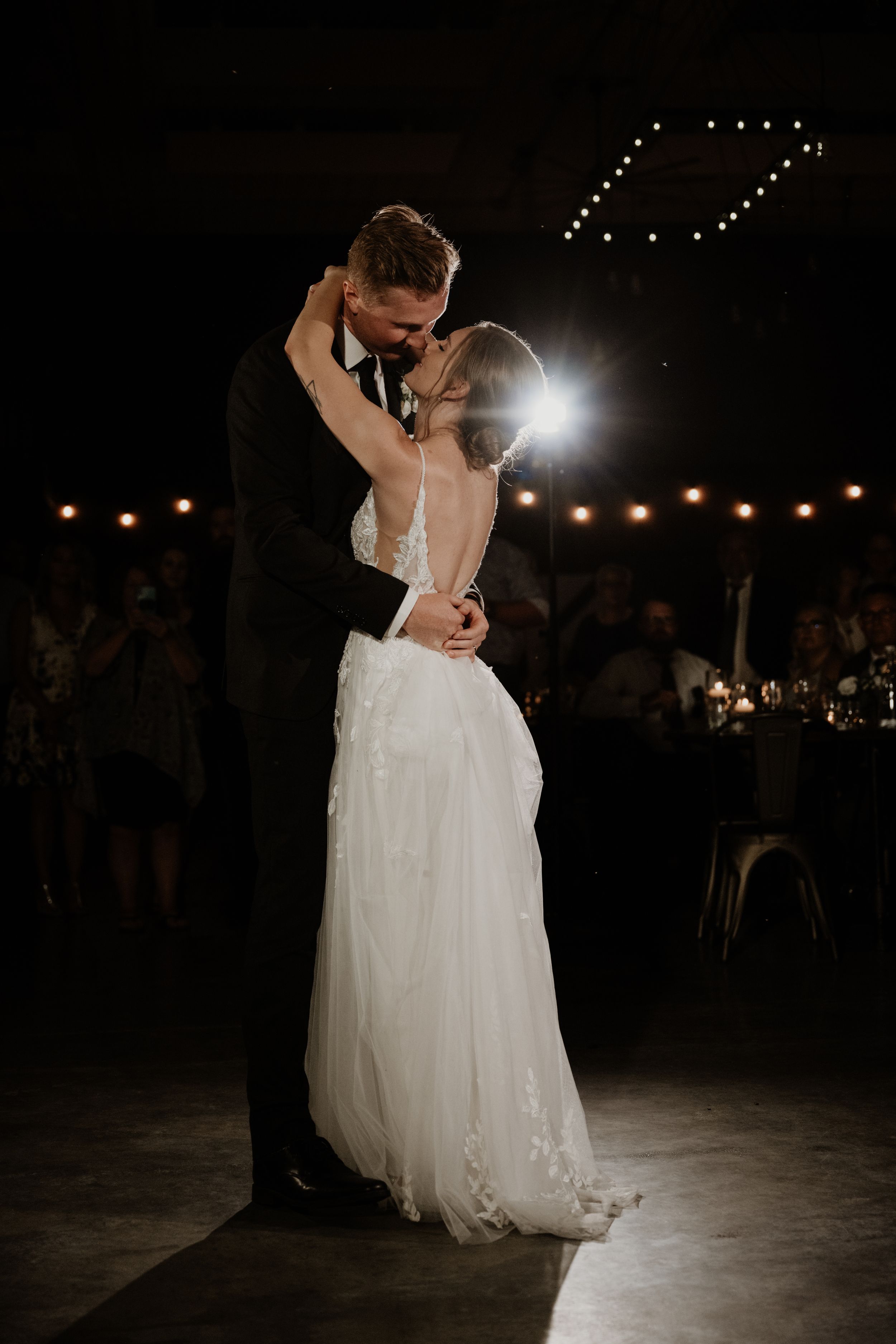 Romantic first dance at a wedding reception with dramatic lighting and white string lights glowing in the background.