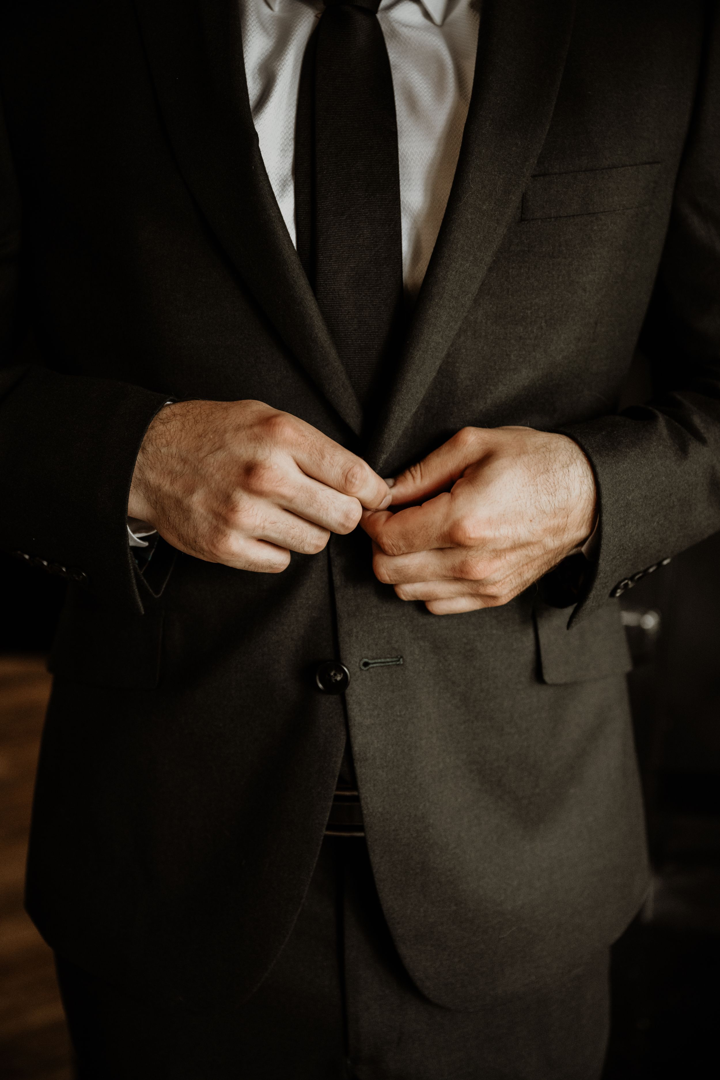 Hands buttoning up a sophisticated dark suit jacket with a light colored tie, captured in moody lighting.