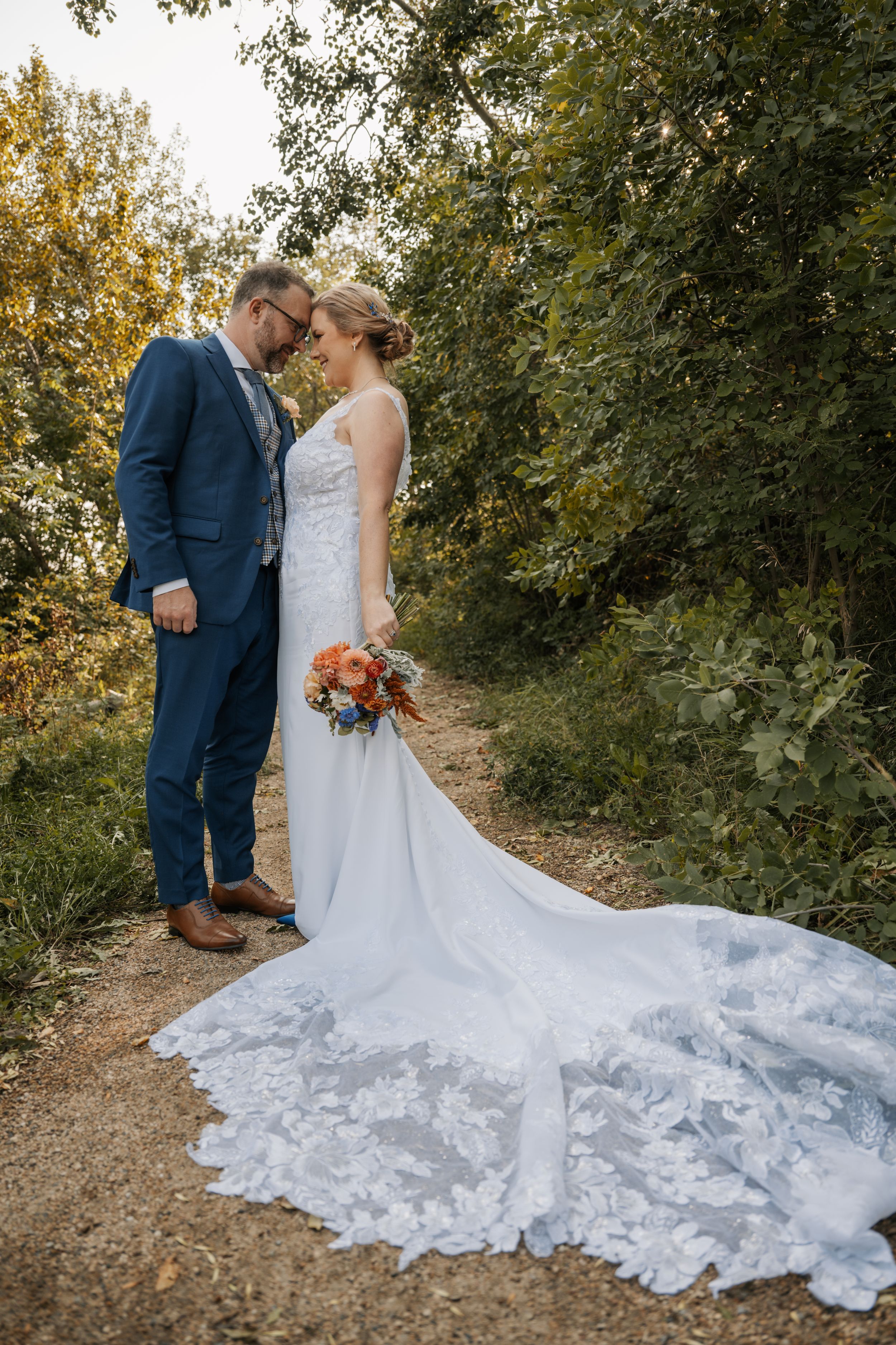 Wedding couple stands on autumn path with elegant white lace gown and navy suit as sunlight filters through trees.
