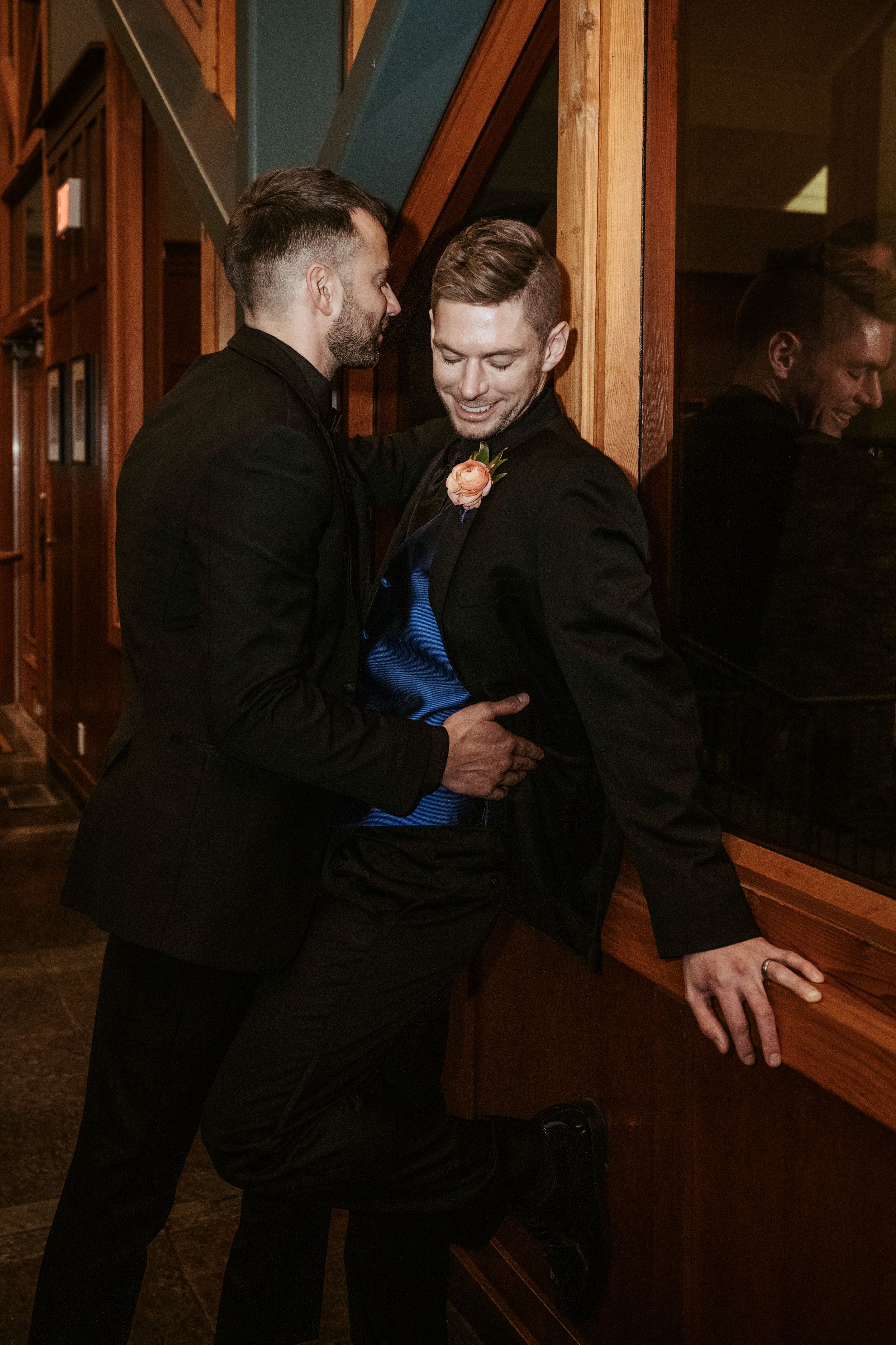 A couple in formal black attire interact in a dimly lit wooden hallway with mirrored reflections visible.
