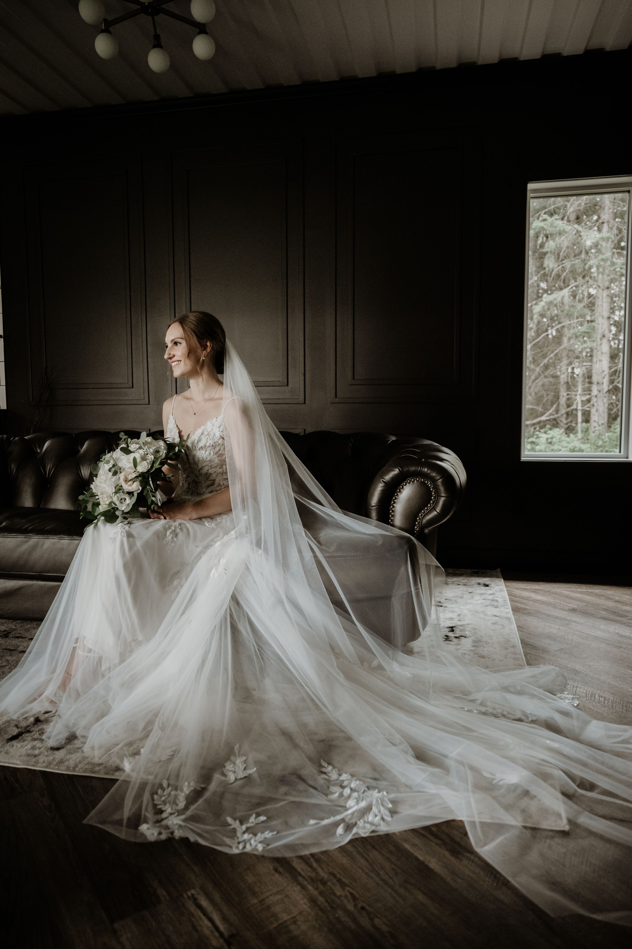 A bride in a flowing white wedding gown with long veil sits elegantly on a dark leather couch holding a white bouquet.