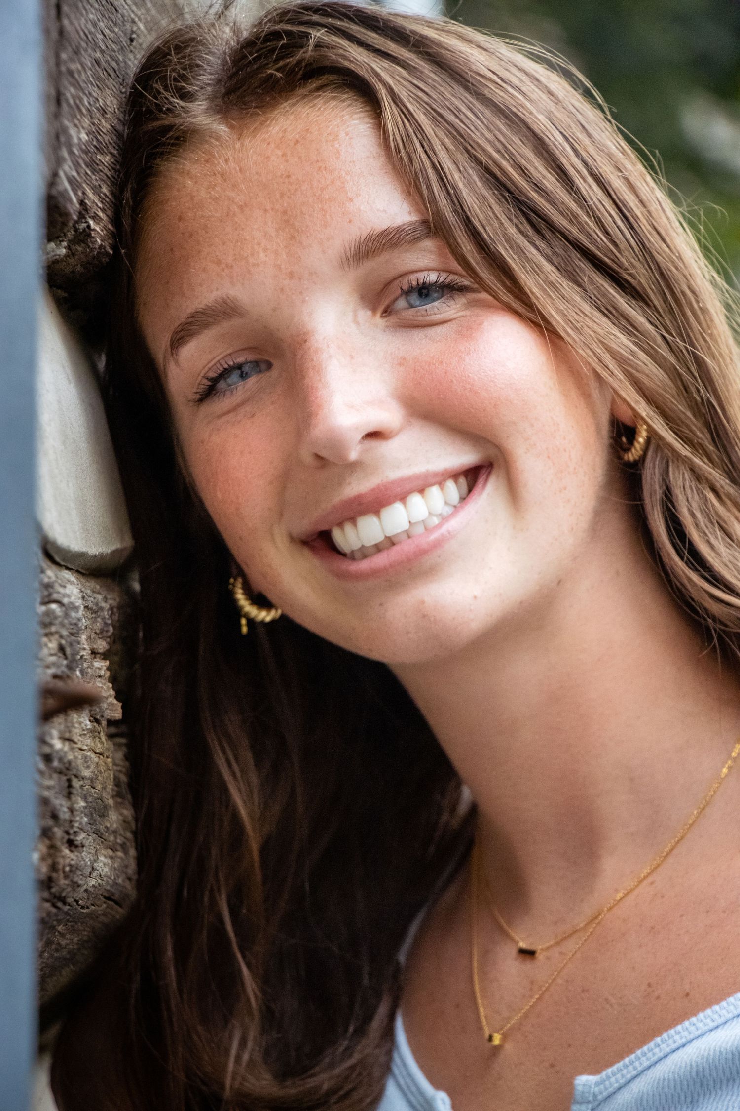 Close-up portrait showing a bright smile and long brown hair against a stone wall background.