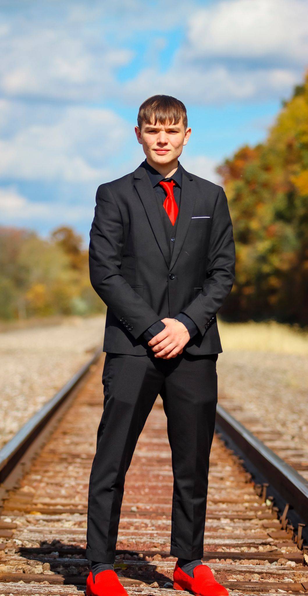 Person in black suit with red tie and shoes standing on railroad tracks in autumn.