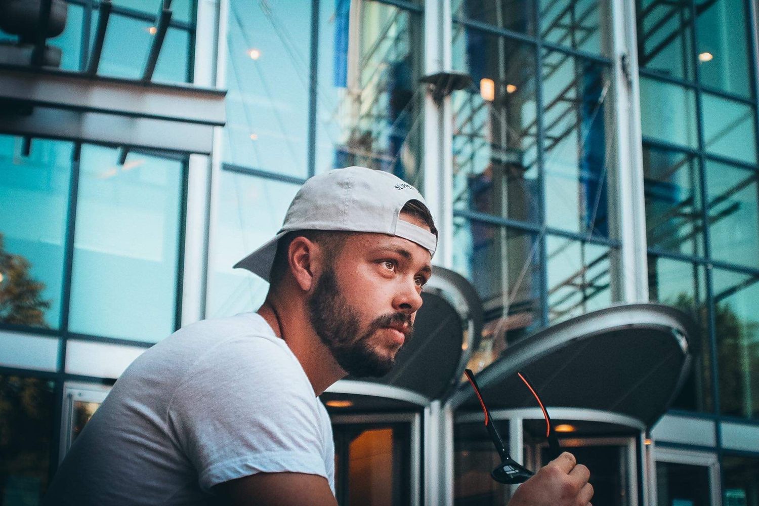 Person in baseball cap and grey shirt sits thoughtfully by modern building.