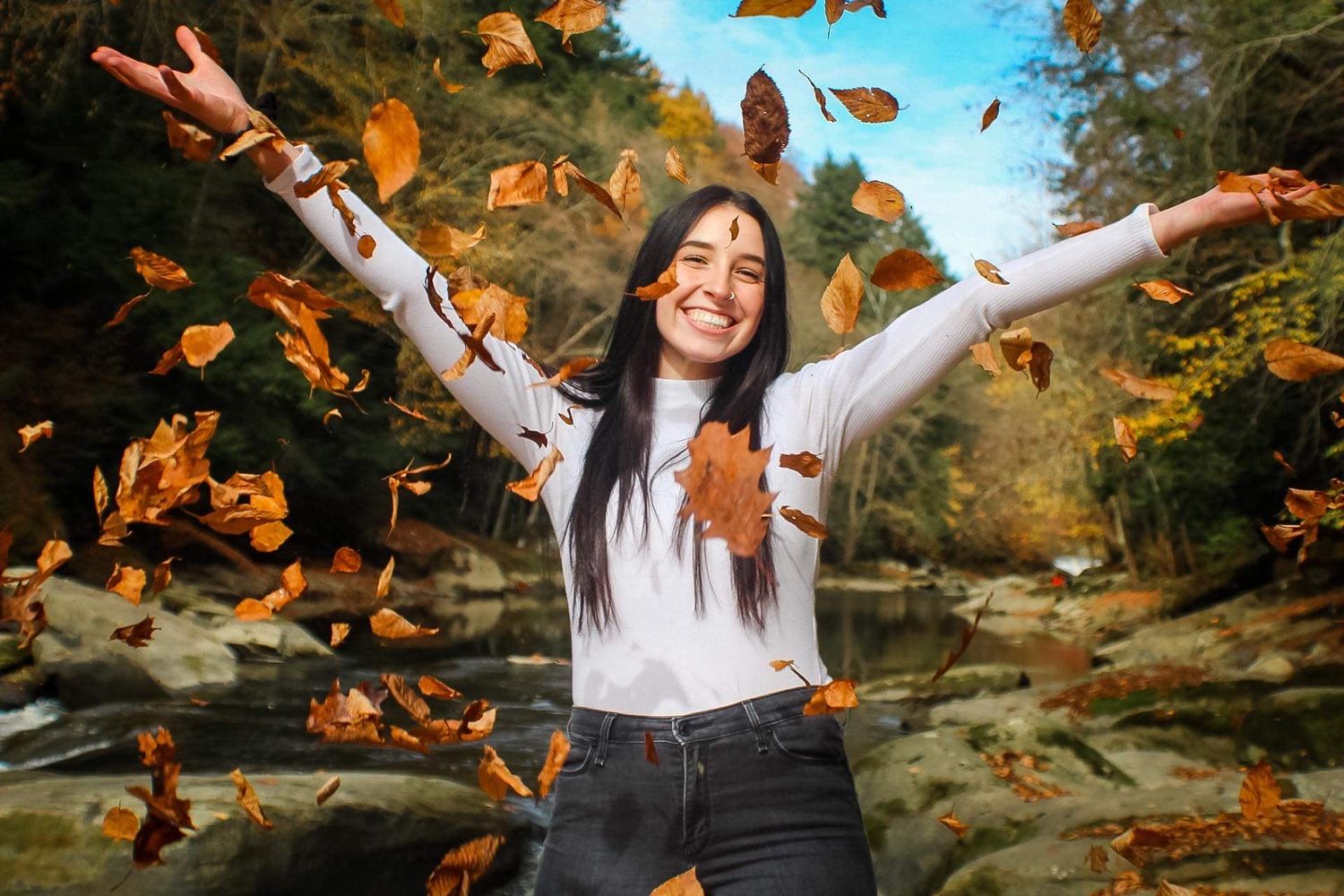 Woman in white sweater joyfully tosses autumn leaves in the air.
