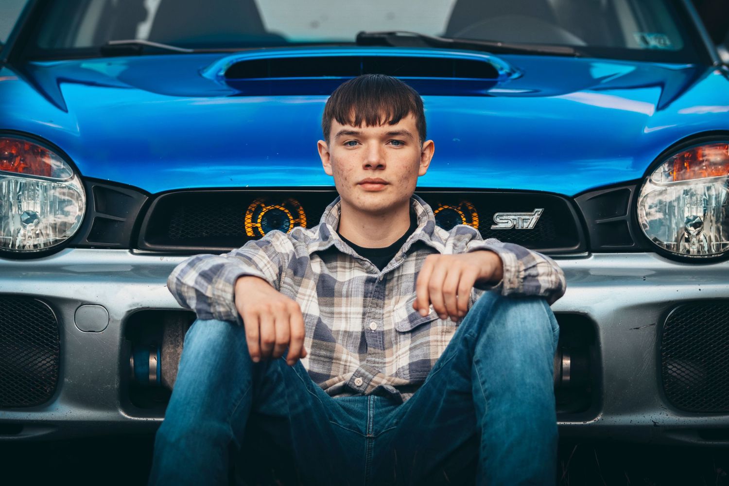 Person sitting on blue car hood wearing plaid shirt and jeans.