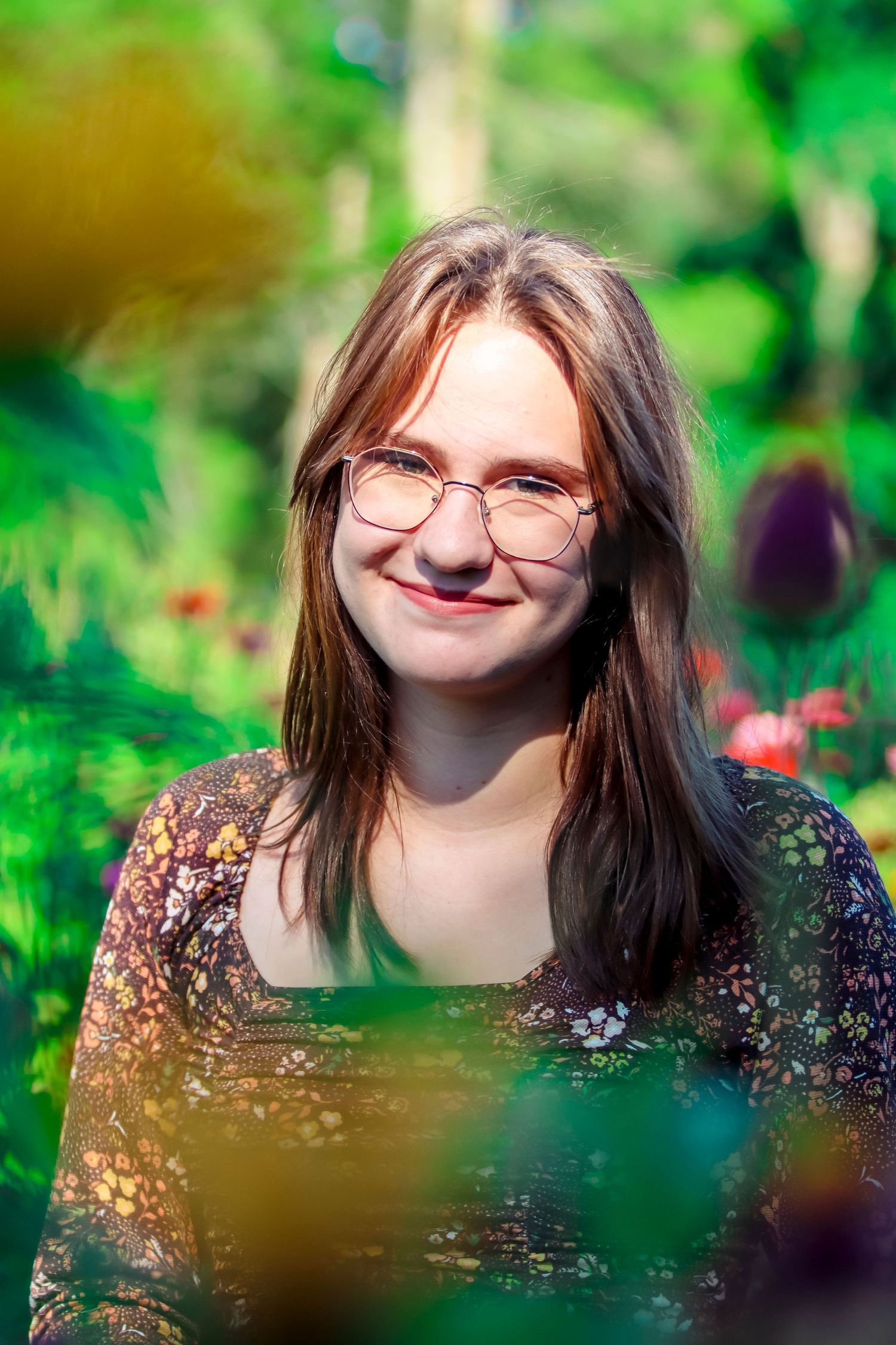 Portrait in a garden with blurred floral bokeh background wearing glasses and patterned top.