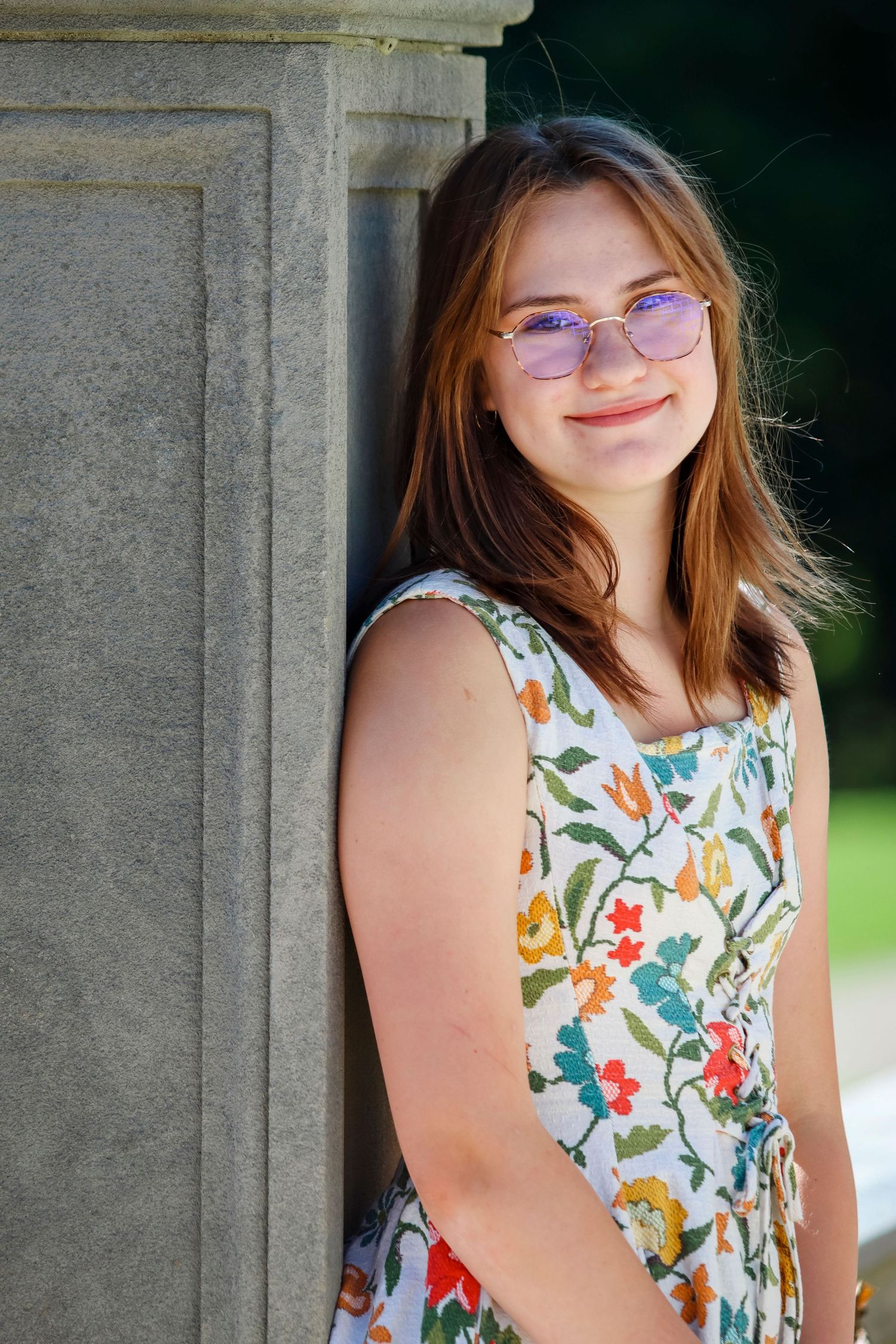 Portrait next to stone column wearing floral summer dress and glasses in natural lighting.