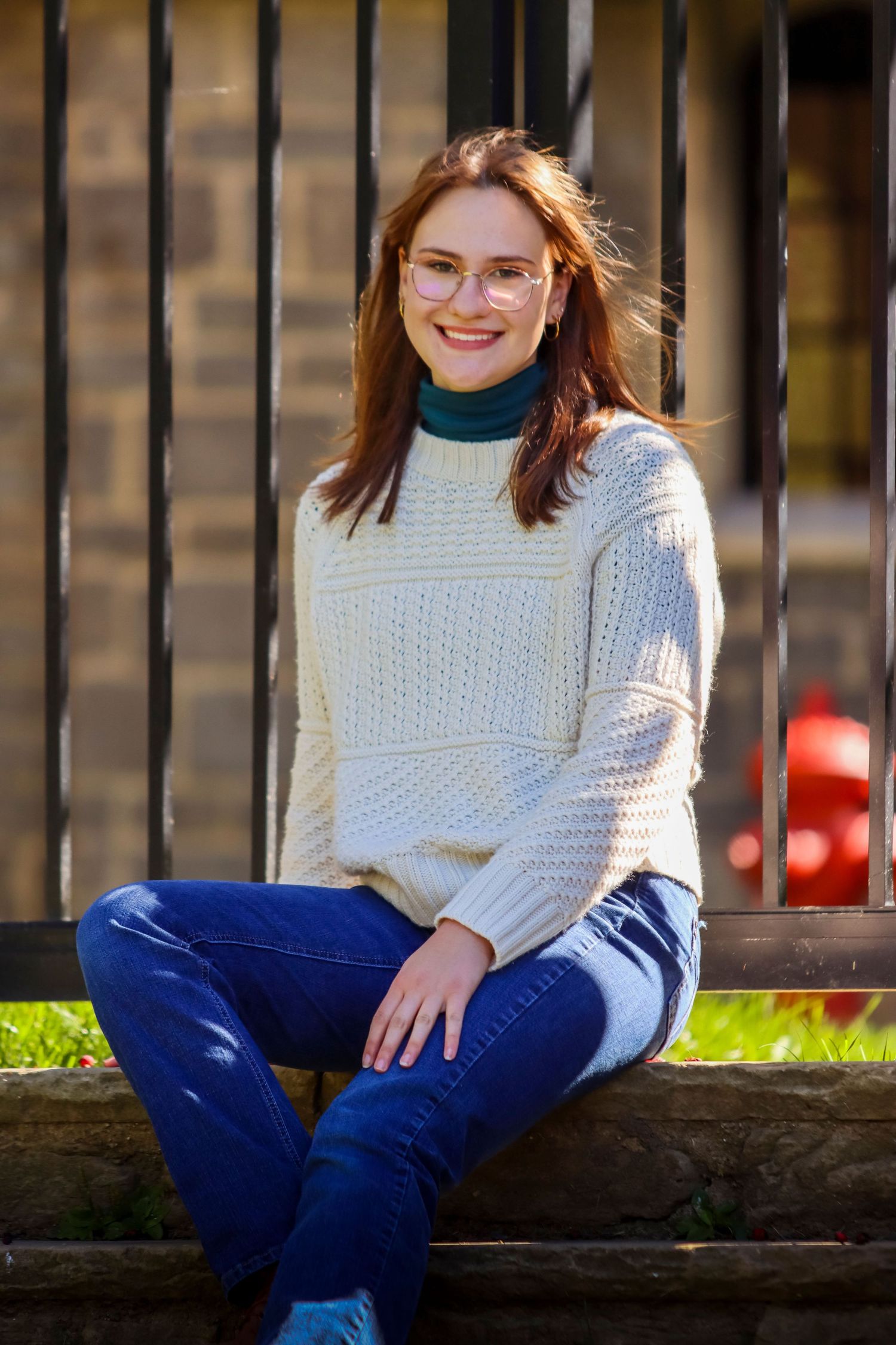 Person wearing glasses and white knit sweater sitting casually on stone ledge outdoors.