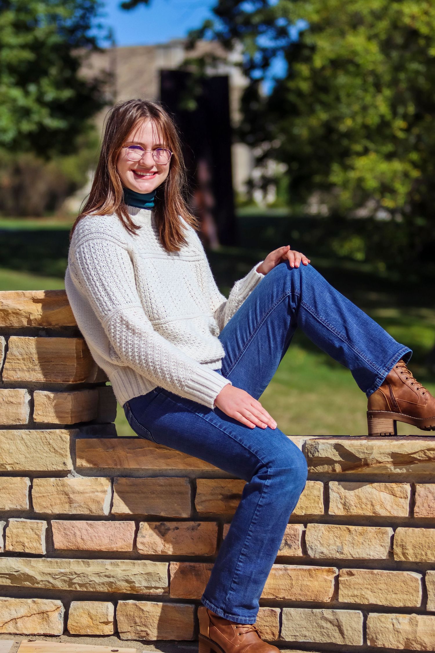 Casual portrait on stone wall wearing white sweater and jeans in sunny outdoor setting.