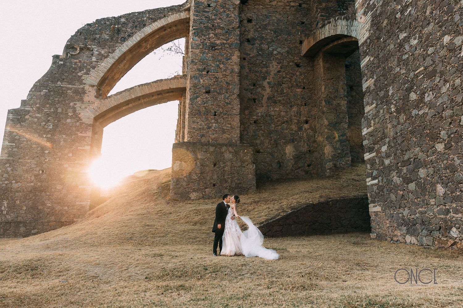 A dramatic wedding photo silhouetted against ancient stone arches at sunset.