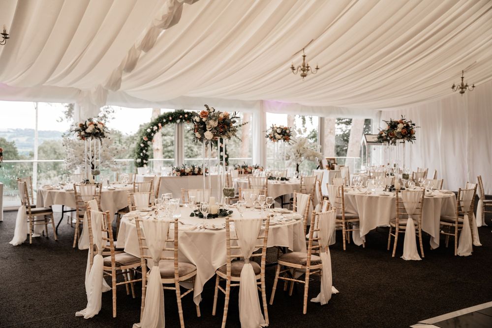 Elegant wedding reception setup in white draped marquee with floral arches and chandelier lighting.