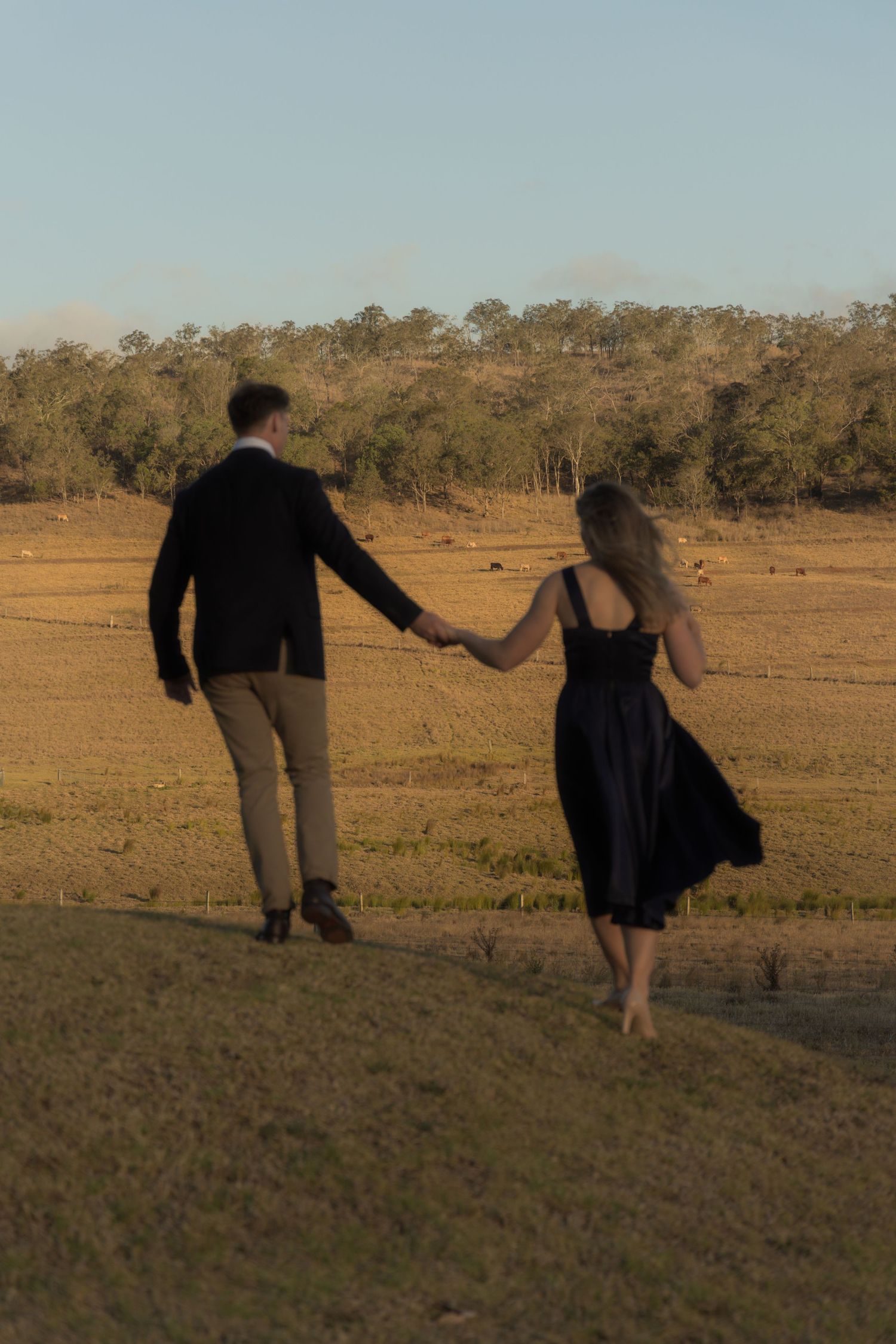 A couple holds hands while walking through a scenic rural field at sunset.