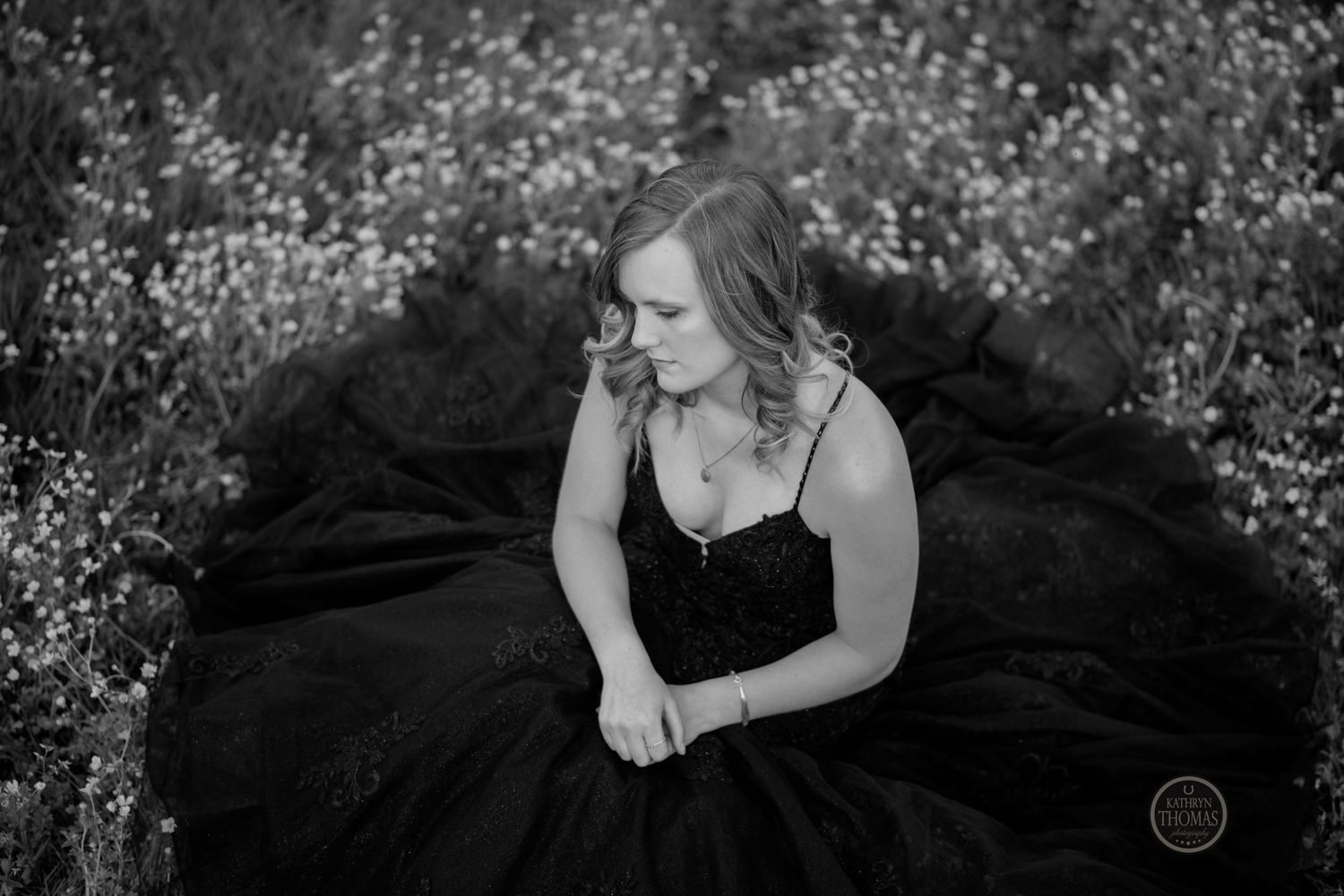 Black and white portrait of woman in elegant ball gown sitting among delicate wildflowers.