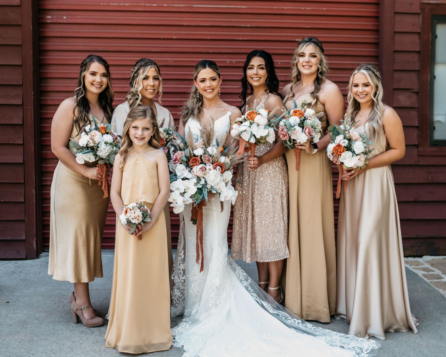 Wedding party in champagne-colored dresses poses with bouquets against a red barn wall.