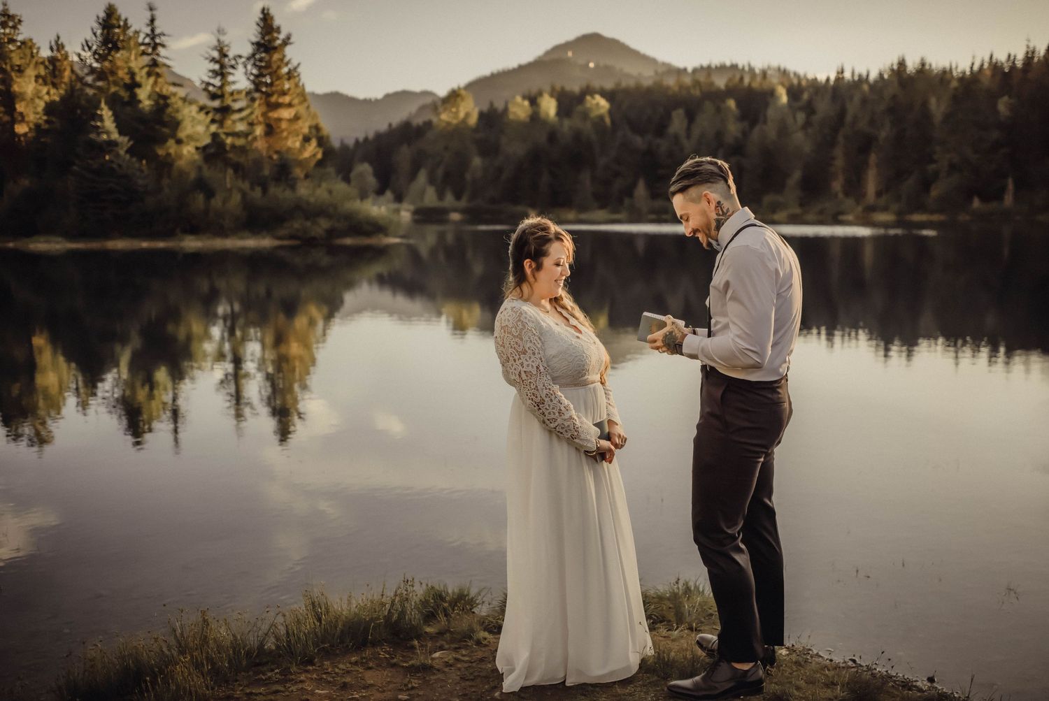 Bride and groom standing by a tranquil lake with forest and mountain backdrop, showcasing destination wedding photograph