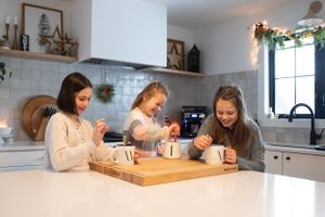 Three people gather around a wooden cutting board on a white kitchen counter.
