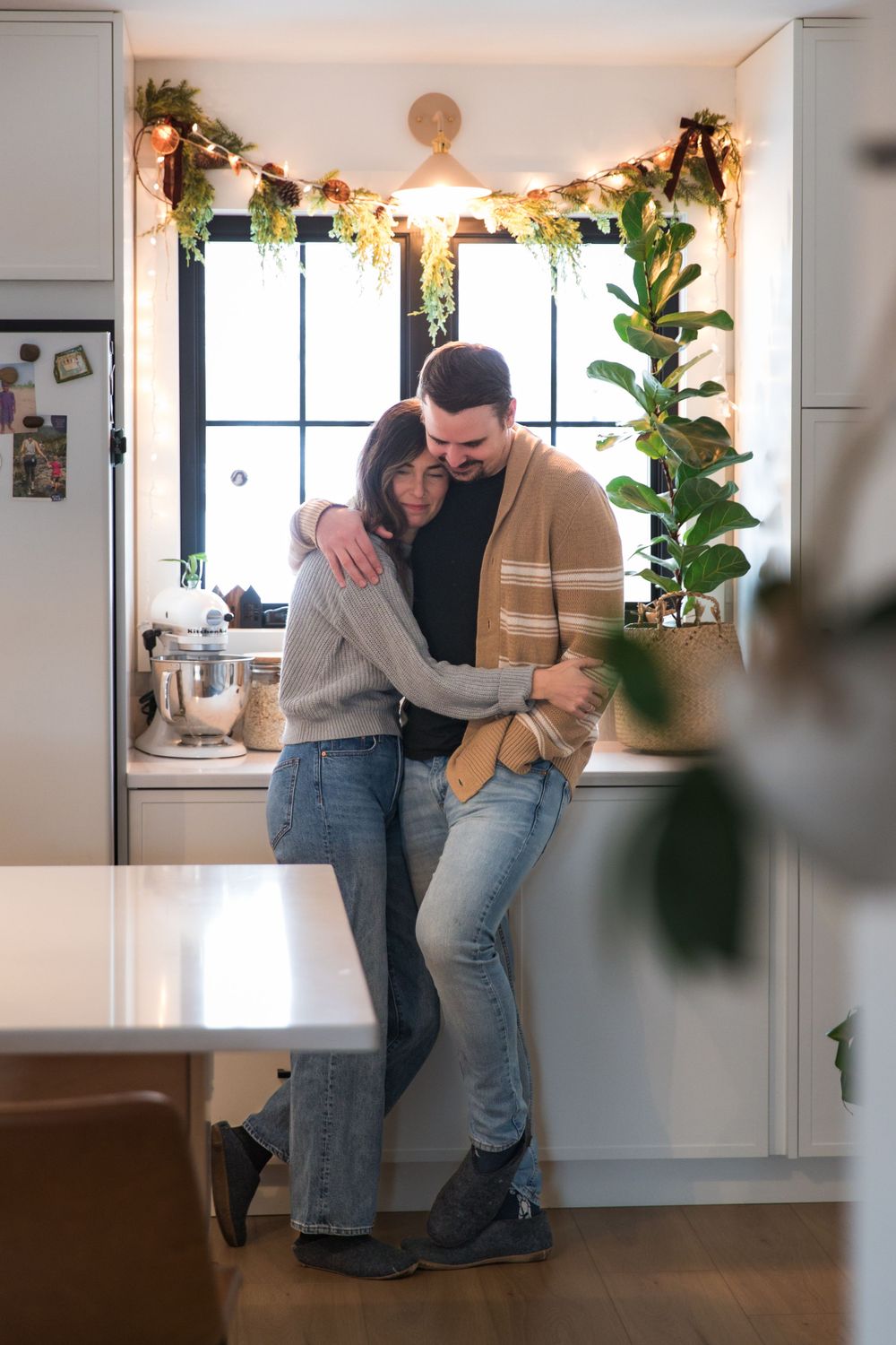 A couple embraces in their cozy kitchen decorated with holiday greenery and lights.