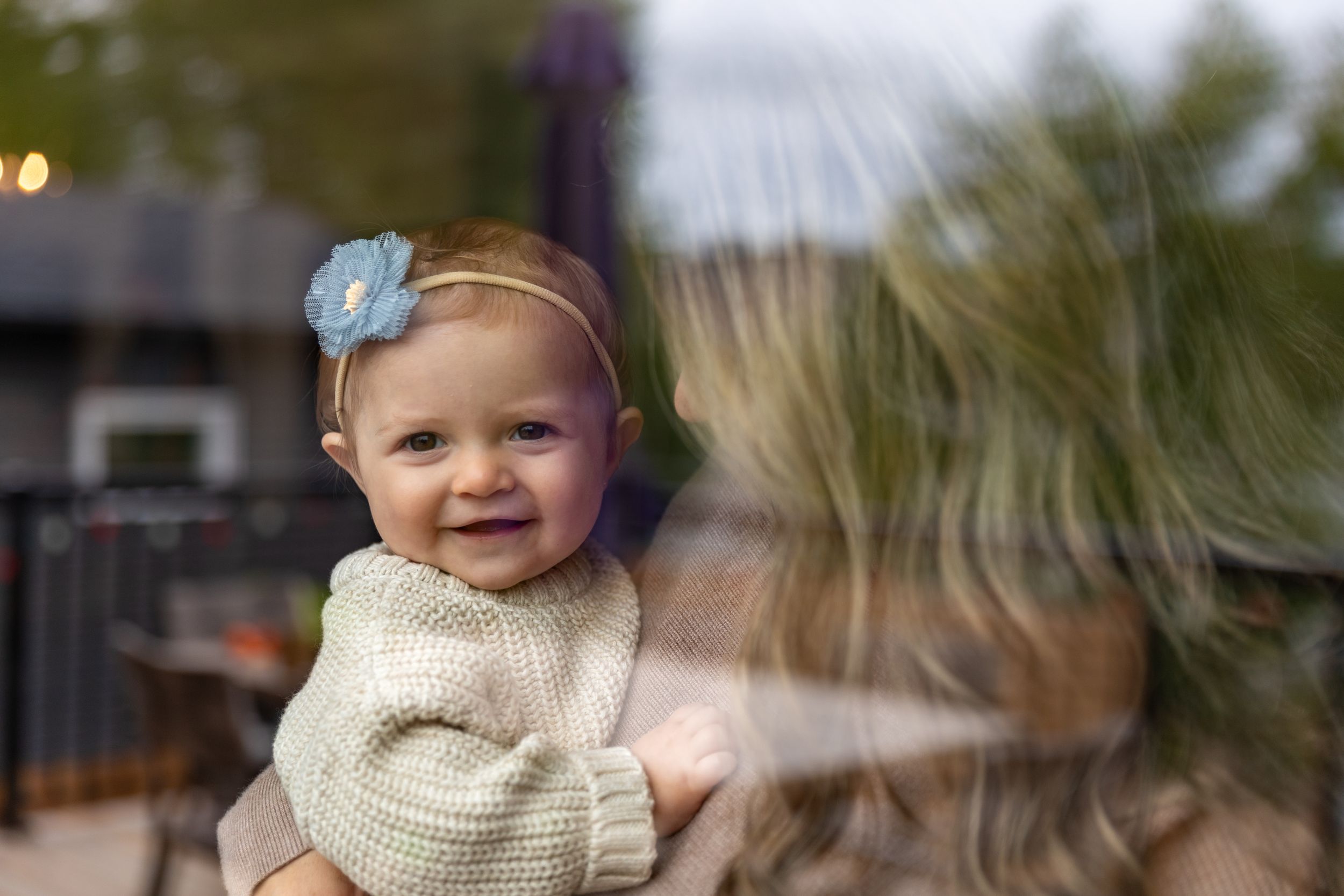 Baby wearing cream sweater and blue headband smiling through window reflection.