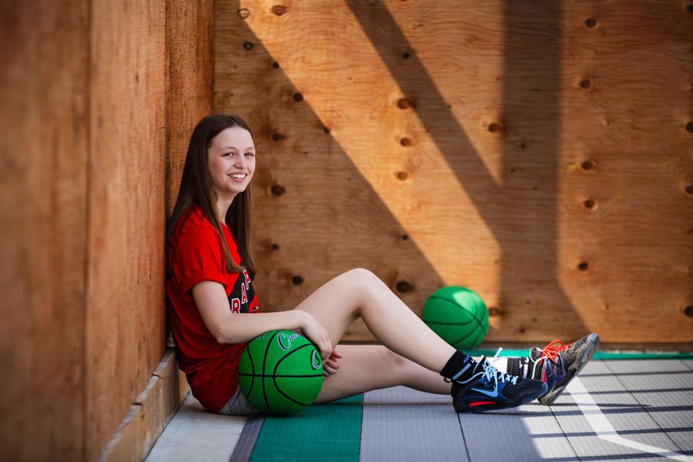 Athletic person sits in sunlit corner of indoor climbing gym wearing red shirt and green shorts.