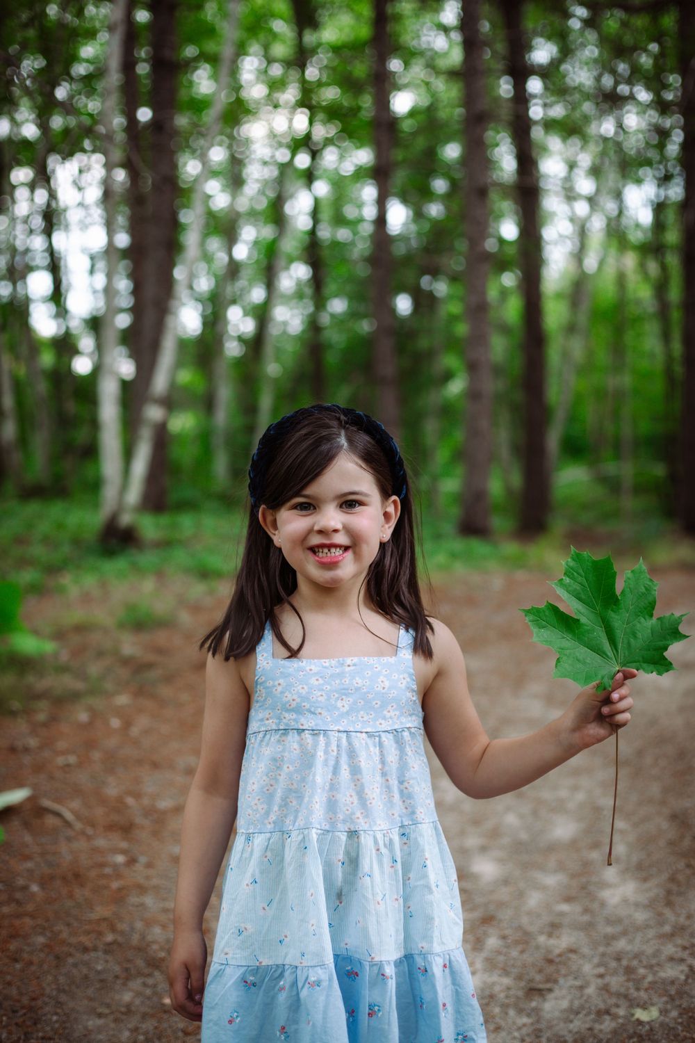 A young girl in a light blue summer dress holds up a large green leaf while standing in a peaceful forest.