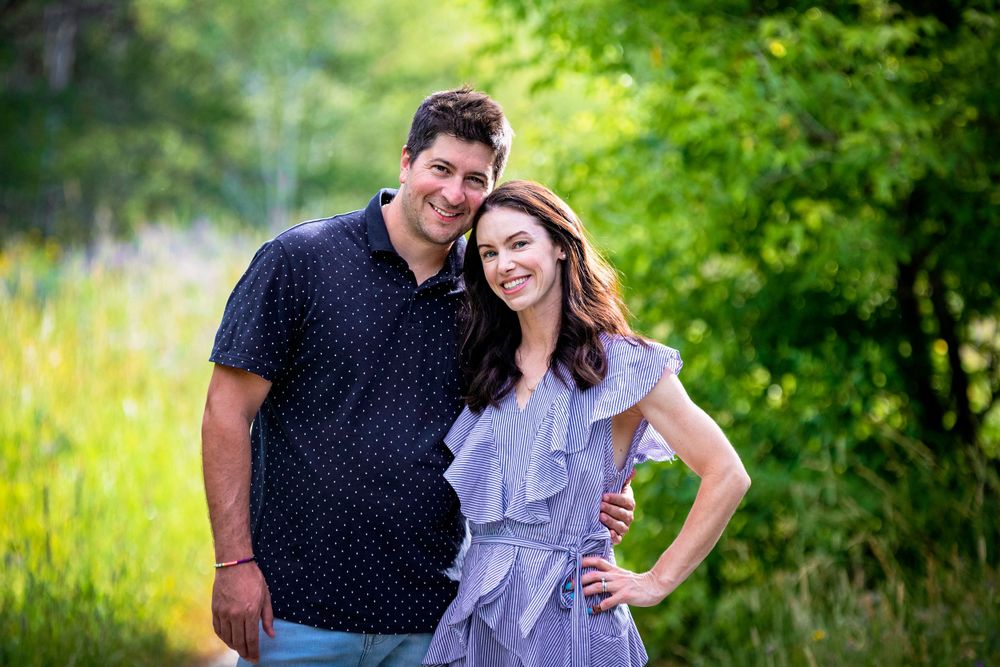 A couple poses outdoors with lush green foliage in the background.