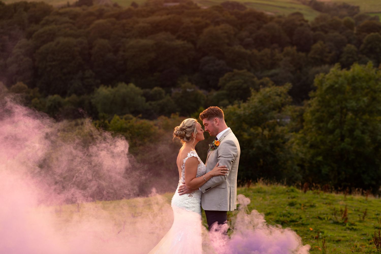 Wedding couple embraces surrounded by pink smoke in countryside setting.