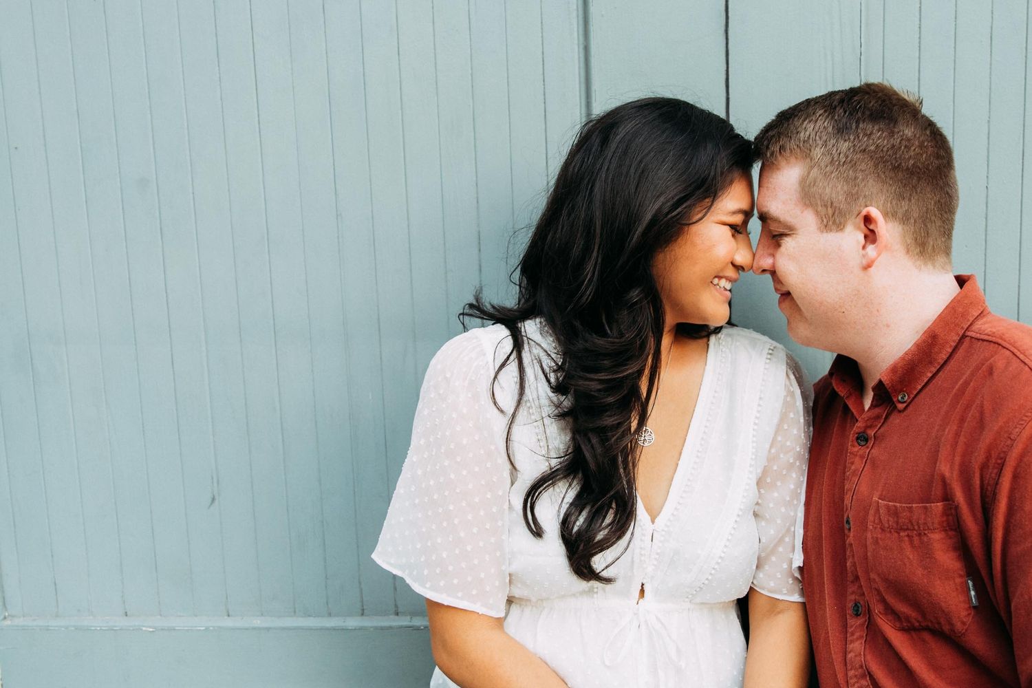 Bi-racial couple sits against blue wall for summer engagement portraits in San Jose.