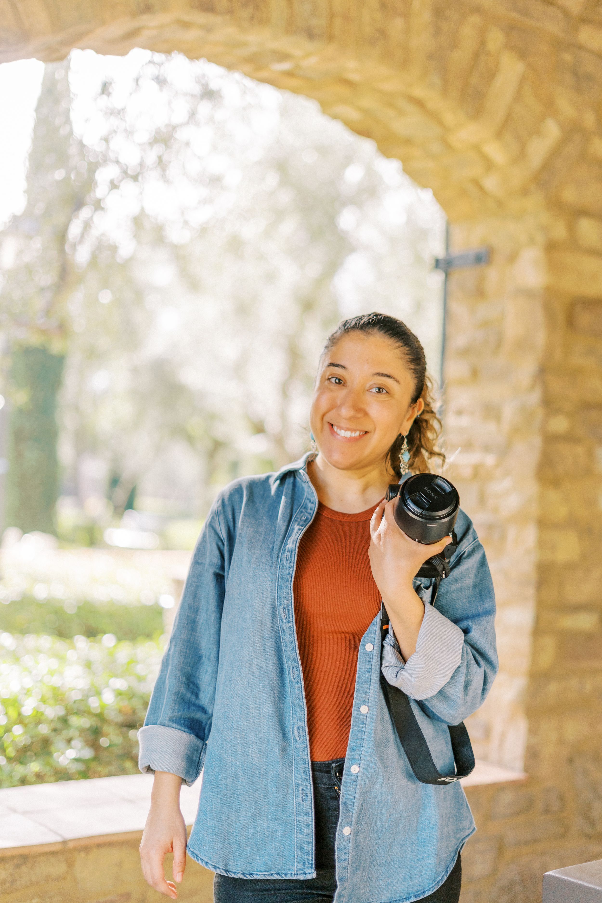 Professional photographer in denim shirt holding camera under stone archway in natural light.