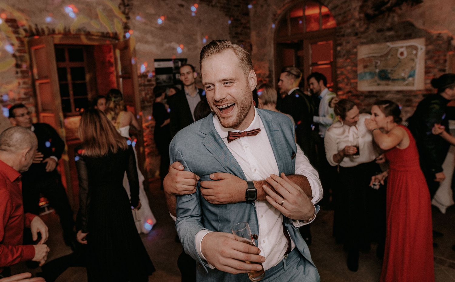 Guest in a grey suit and bowtie dances joyfully at an indoor wedding reception.