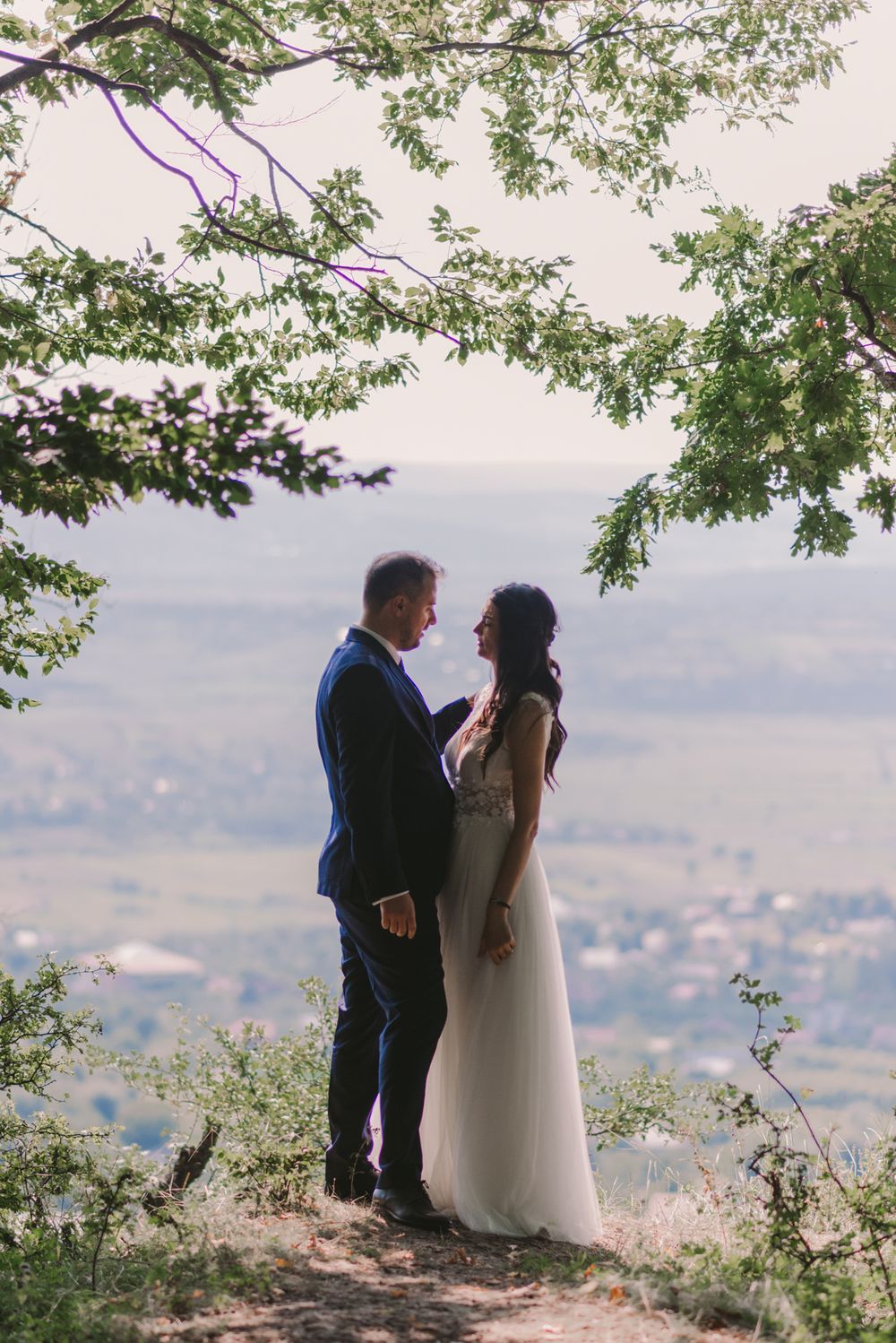 Wedding couple stands on hilltop overlook sharing intimate moment with city views below.