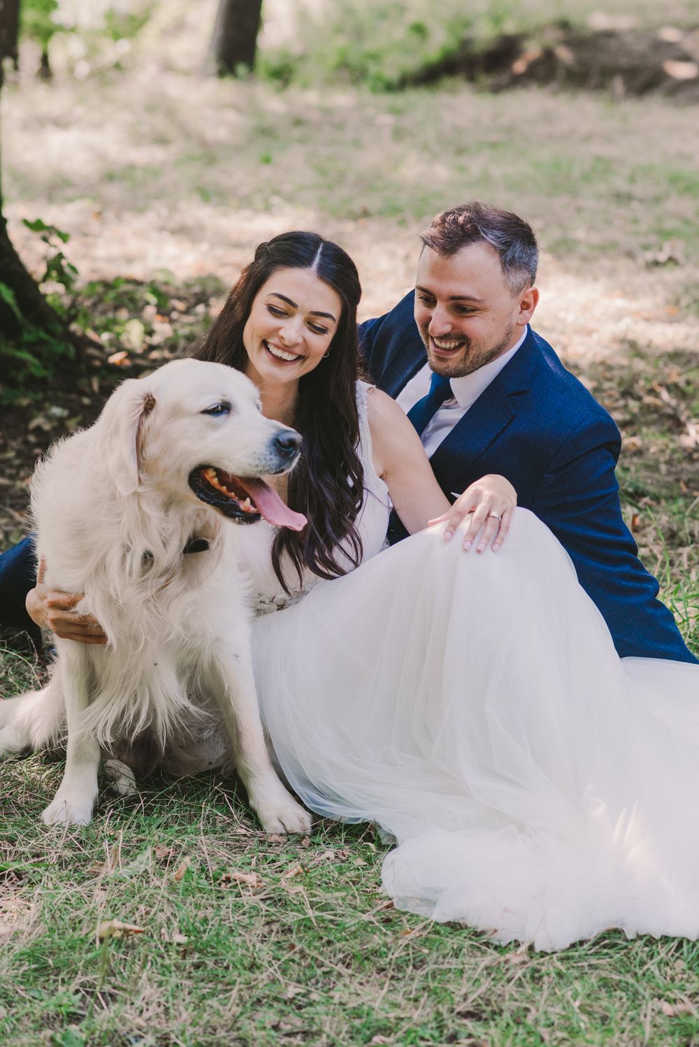 Newlyweds cuddle with their white dog on grass during outdoor wedding photos.