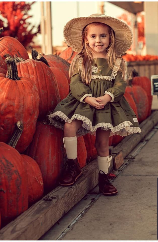 Child in vintage dress and sun hat posing with large orange pumpkins on wooden platform.
