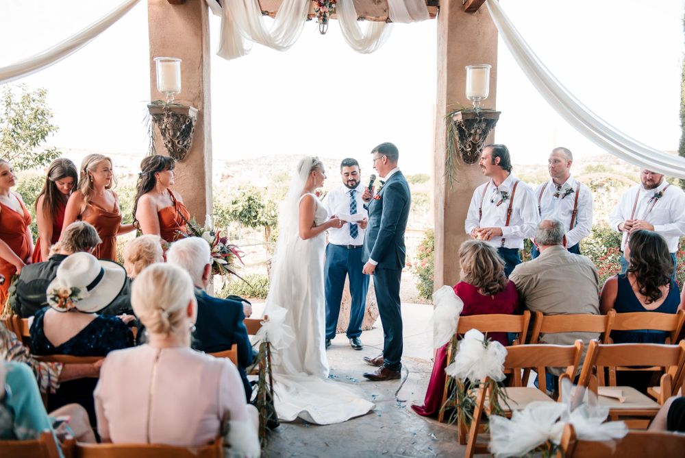 Outdoor wedding ceremony with guests seated on wooden benches under a decorated archway.