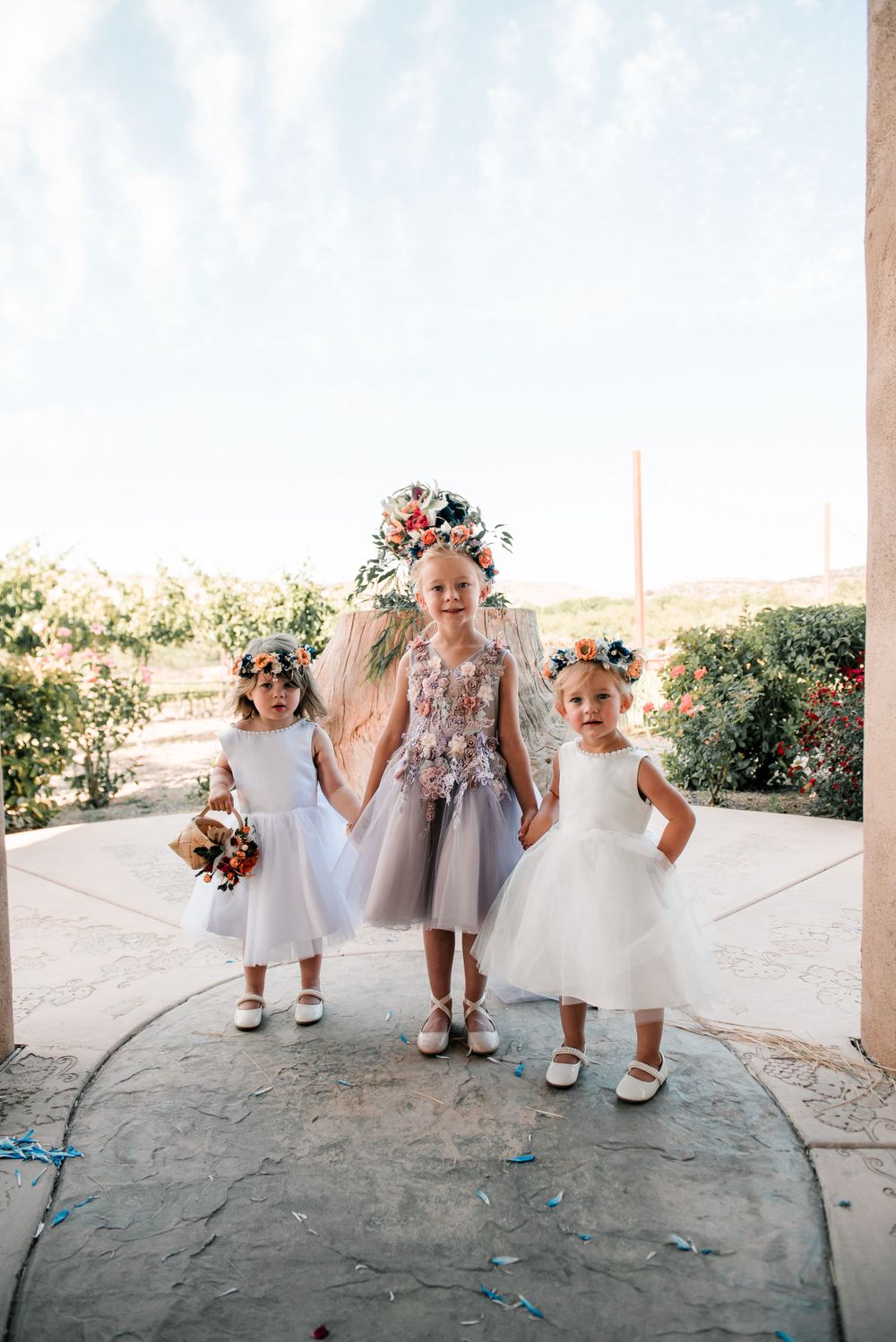 Three young attendants in white dresses posing together at an outdoor wedding venue.