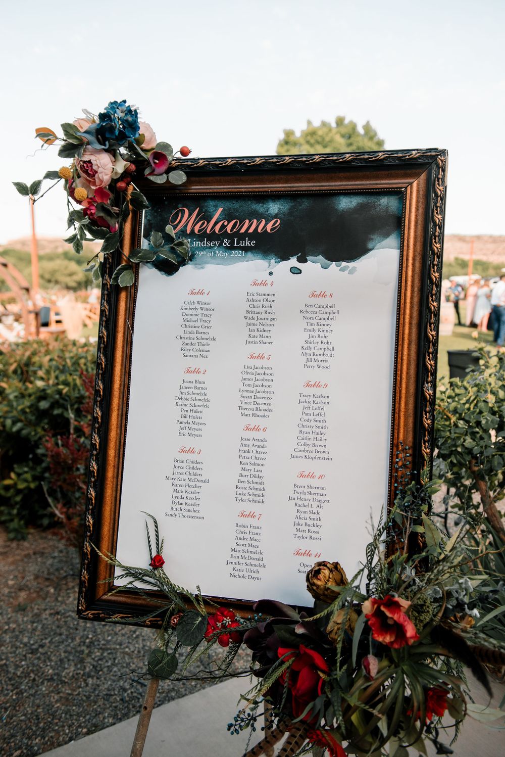 Ornate framed seating chart with floral decorations displaying wedding guest names against a white background.