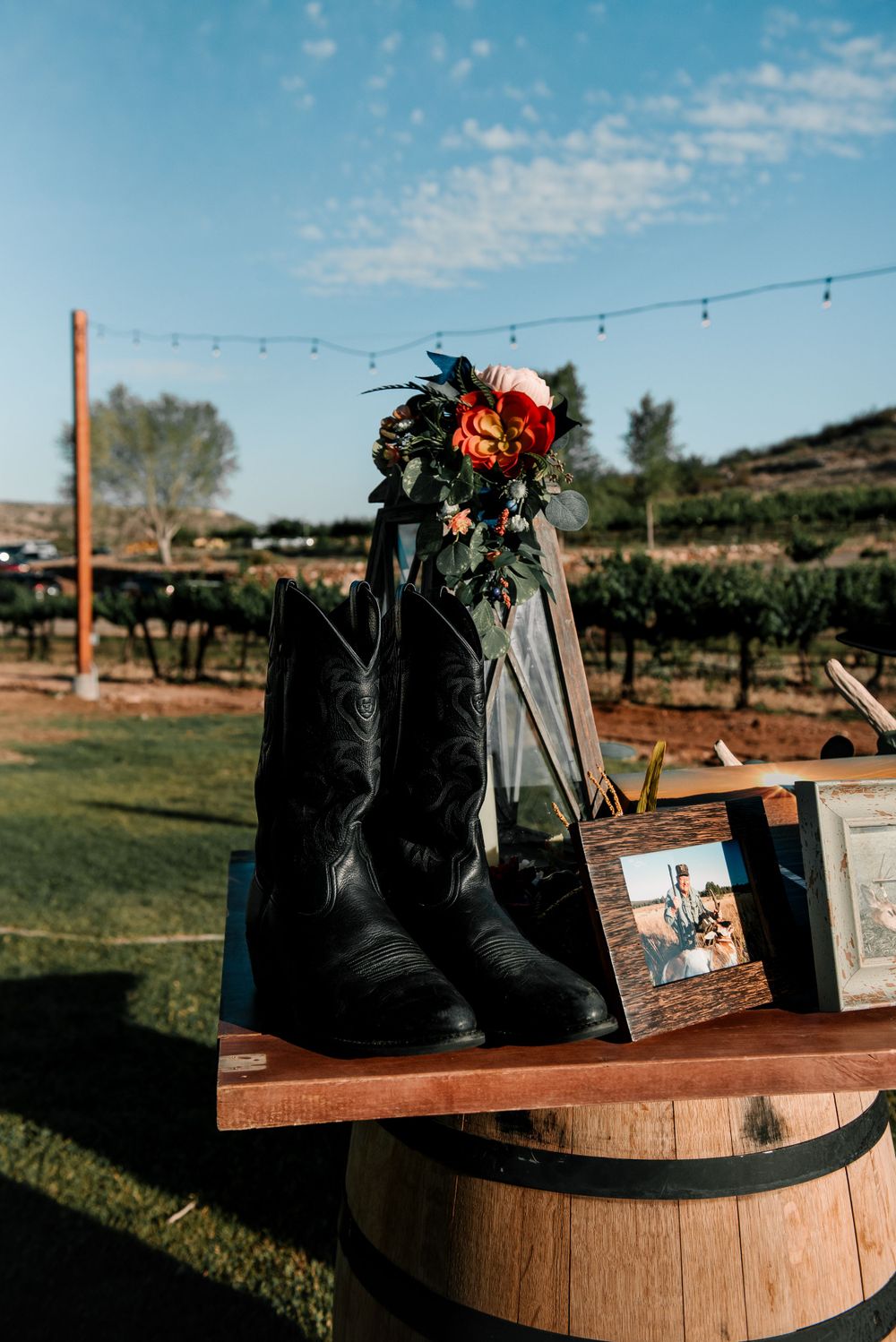 Black boots adorned with flowers displayed on wooden barrels at an outdoor wedding venue.