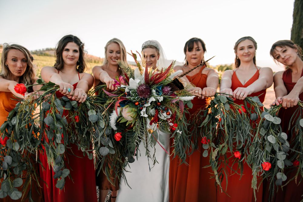 Bridesmaids in burgundy dresses with floral garlands at outdoor wedding venue.