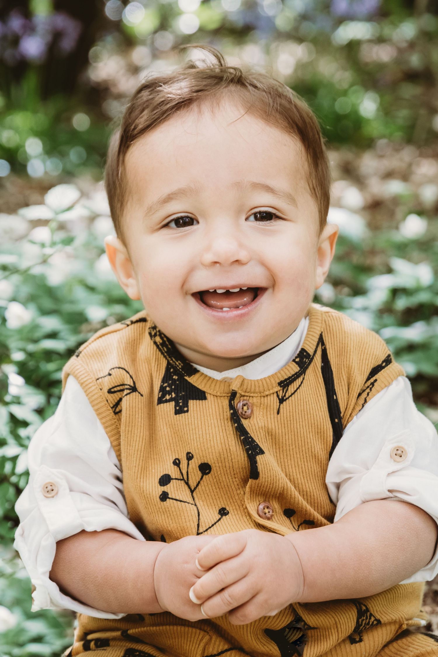 Toddler wearing mustard yellow overalls smiles brightly in woodland setting, Highgate Woods, London.