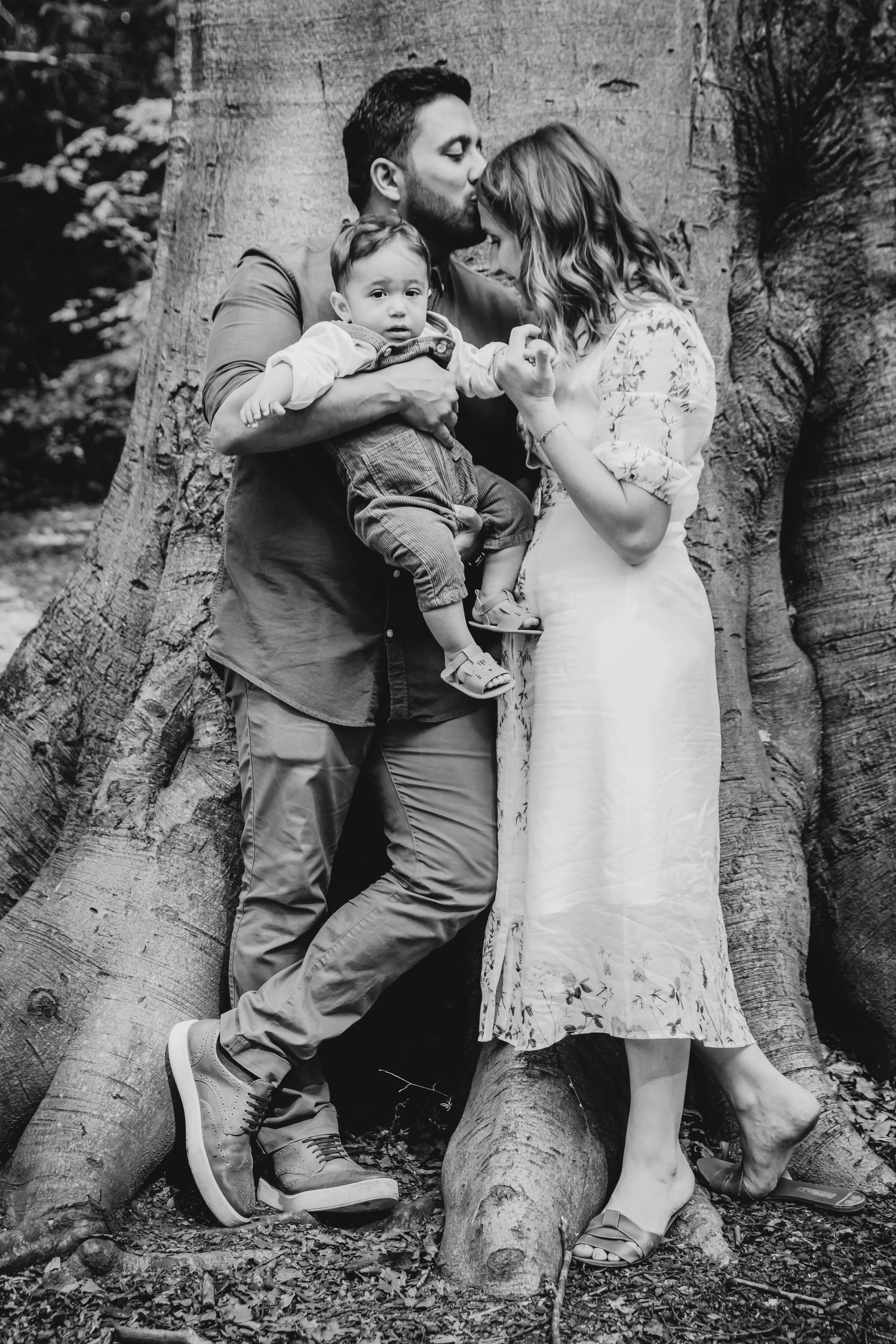 Family shares intimate moment against massive tree trunk in black and white portrait, Highgate Woods, London.