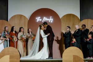 Bride and groom kissing on church stage with neon sign behind, as bridal party cheers.