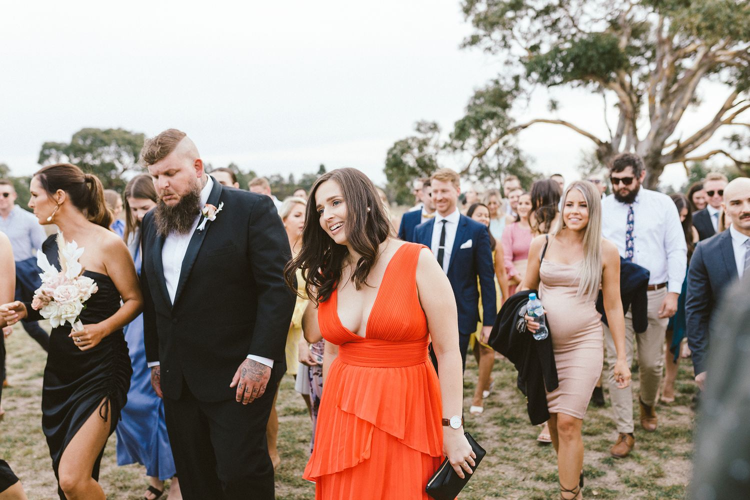 Wedding guests in formal attire walking through an outdoor venue with a large tree in background.