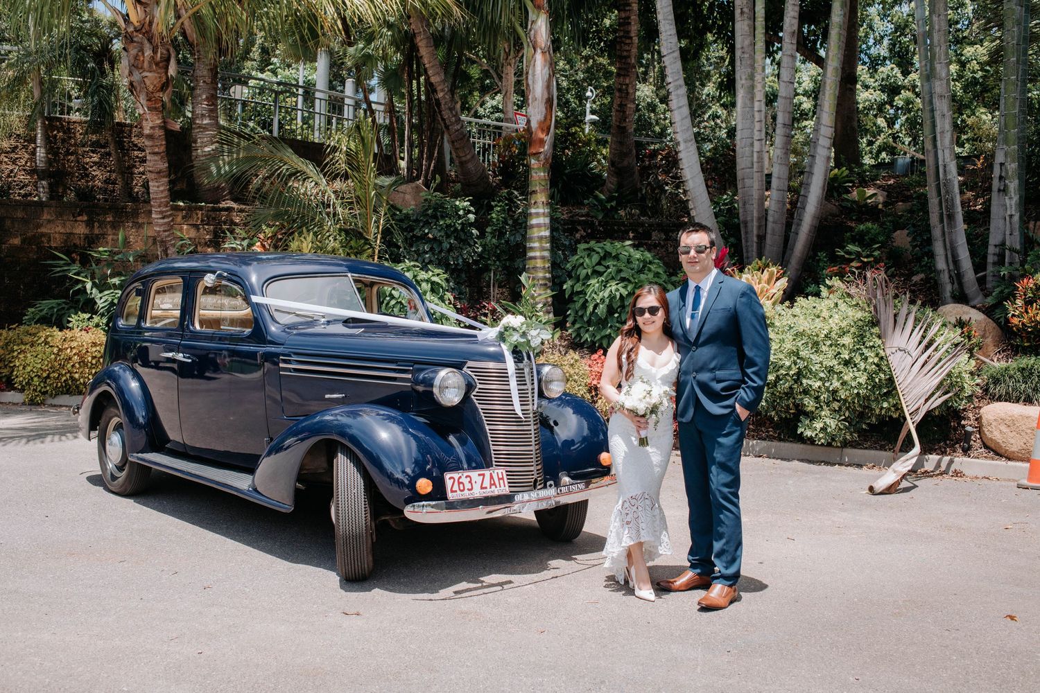 Wedding couple posing with classic vintage blue car in tropical setting.