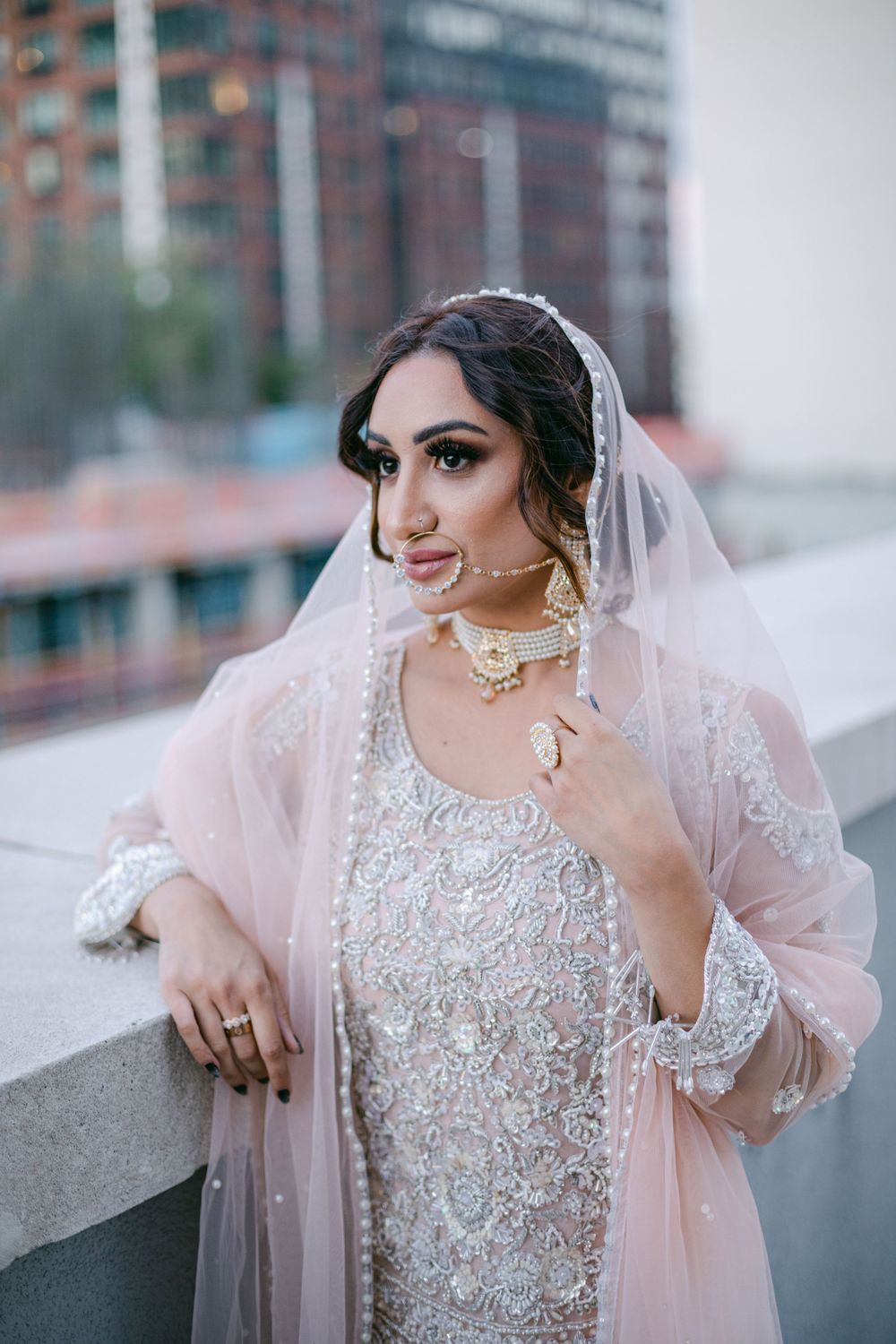 Elegant bridal portrait with pearl jewelry and blush pink veil against city backdrop.
