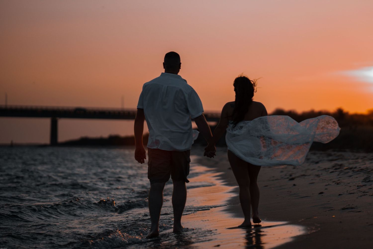 Two silhouettes walk along a beach at sunset with one person carrying a surfboard.