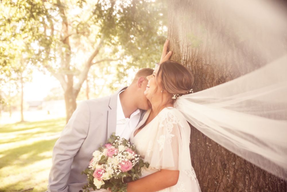 laughing bride holding pink rose bouquet leans on tree while groom smiles at her