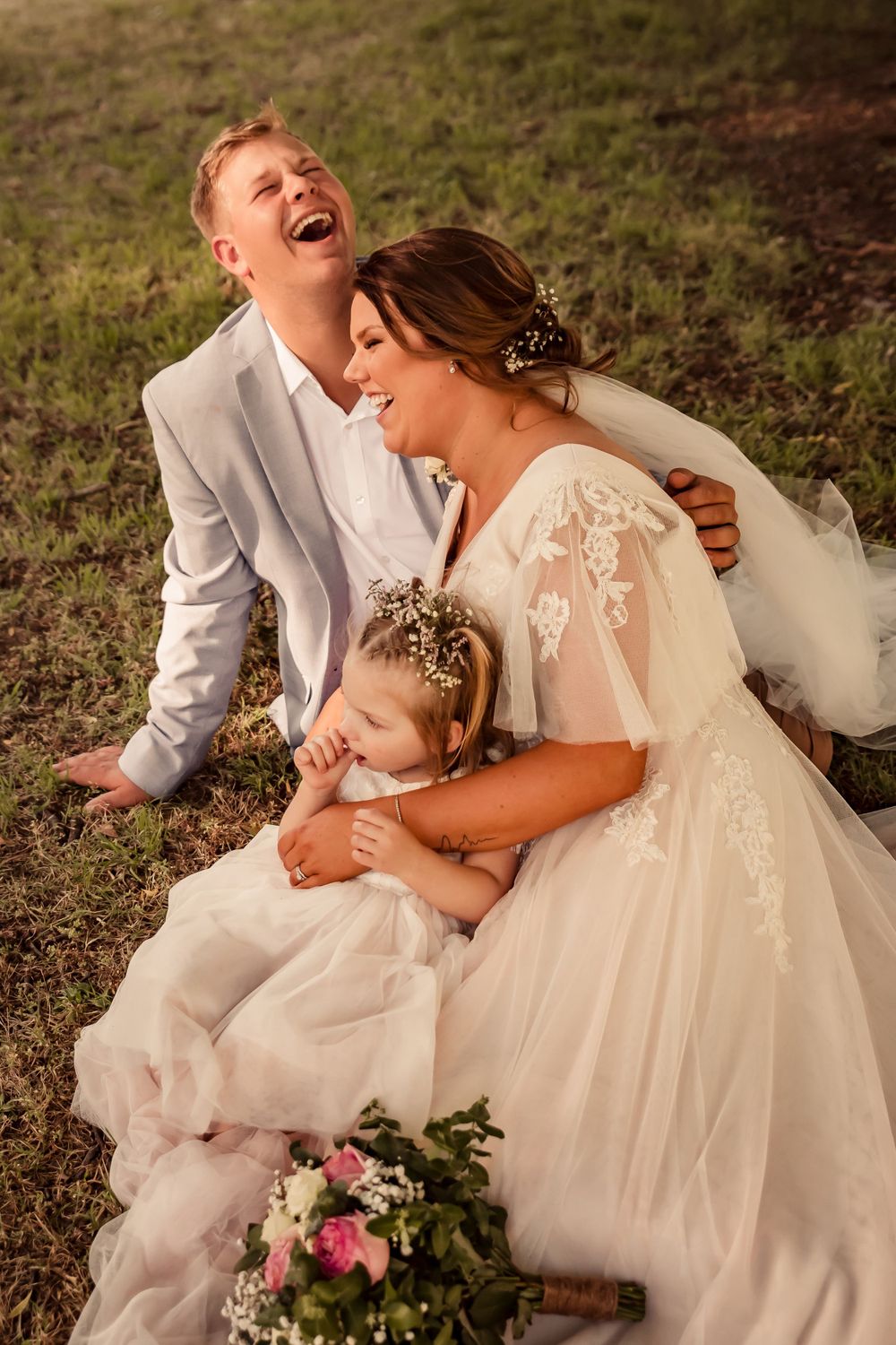 family photo of laughing bride and groom relaxing on grass as they hold their young daughter