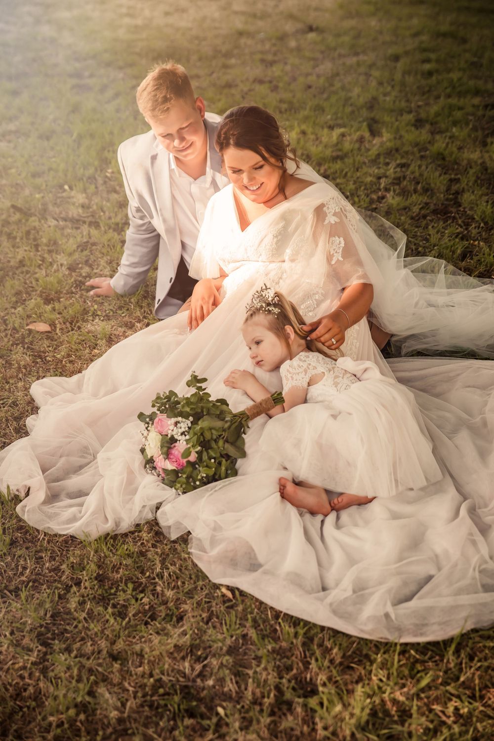 bride and groom reclining on grass and looking at flowergirl who is laying on brides train the sun is shining on them