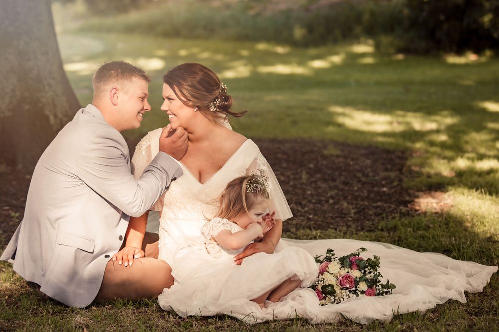 groom embracing bride who is smiling at him as they sit under oak tree.  their daughter is sitting on the brides gown