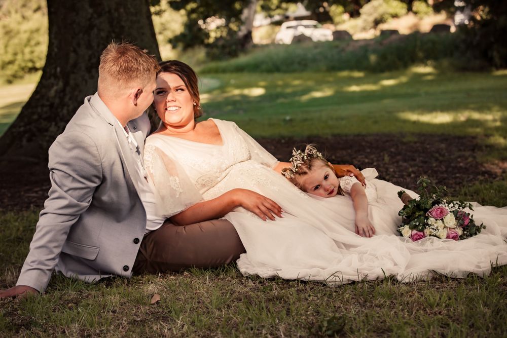 bride laying in grooms lap under large oak tree as their daughter leans on her while holding her bouquet